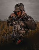 Hunter wearing windproof camo jacket on an open ridgeline with tall grass blowing, prepared for long-range glassing in gusty conditions.