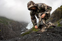 Hunter in camouflage rain gear adjusting gaiters on a mountain slope near a glacier and waterfall in misty weather
