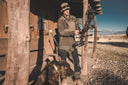 A man wearing Pnuma Outdoors gear stands with a dog outside a rustic cabin in a natural setting.