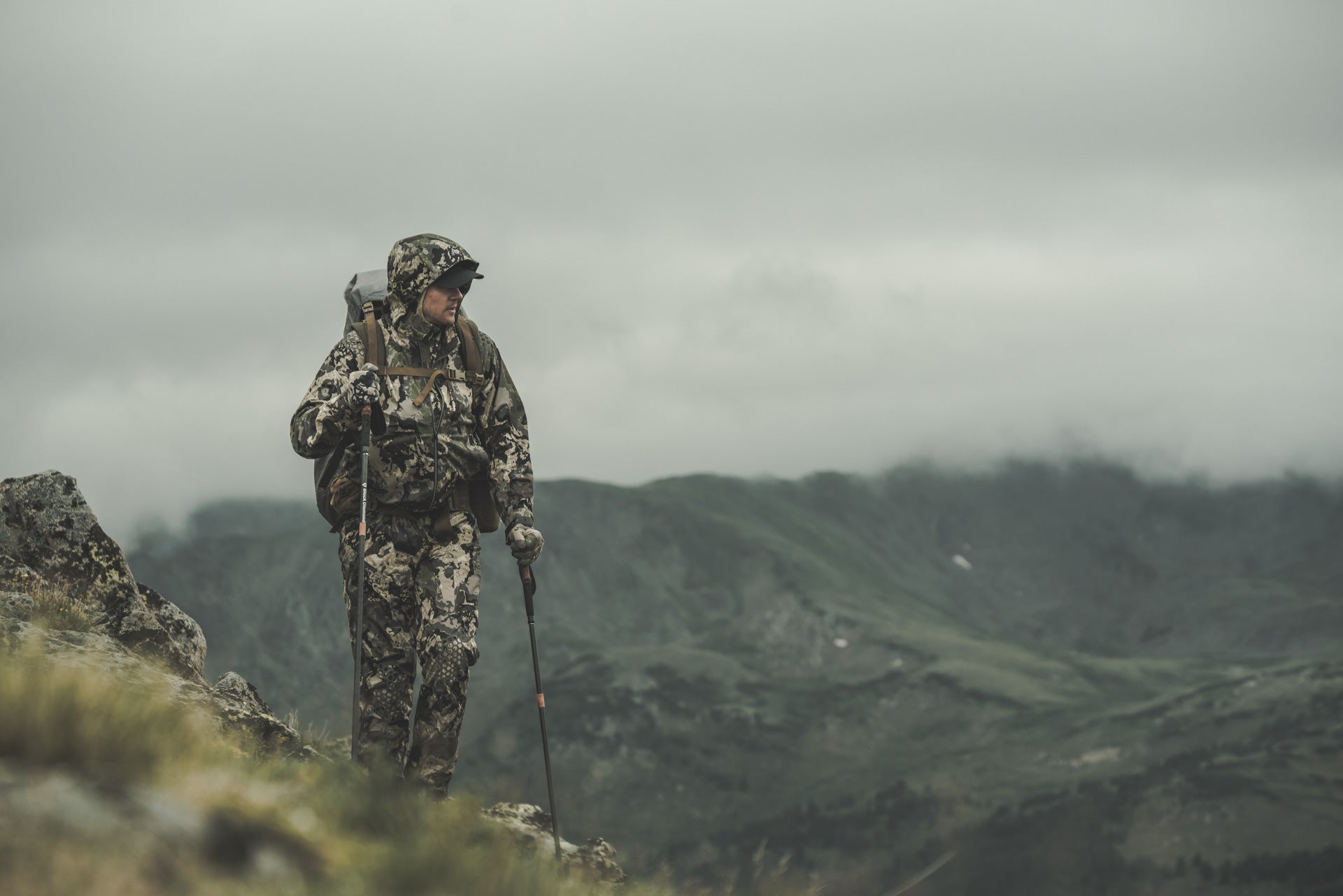 Hunter in full camouflage hiking with trekking poles across a misty mountain landscape.