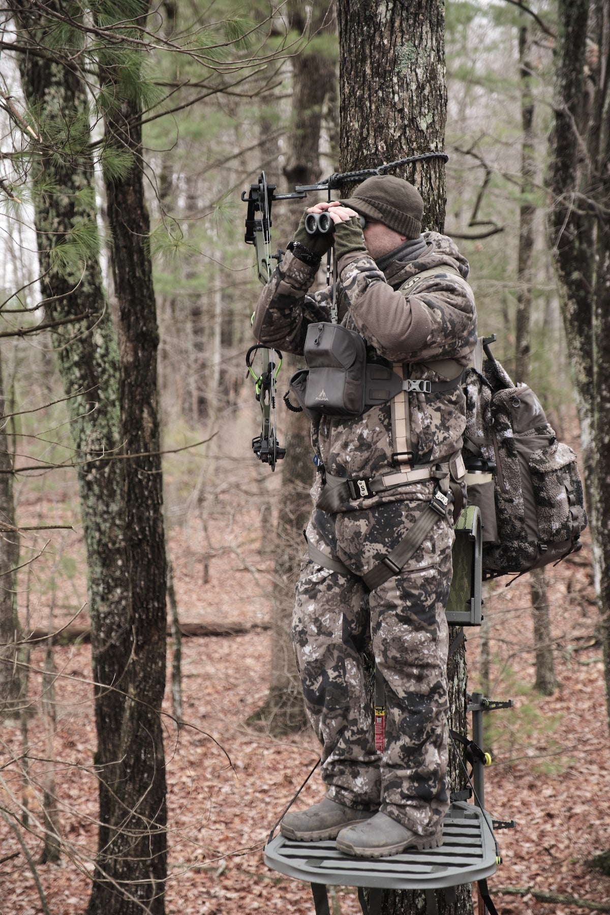 Hunter in full camouflage gear standing on a tree stand, using binoculars to scout in a wooded area.