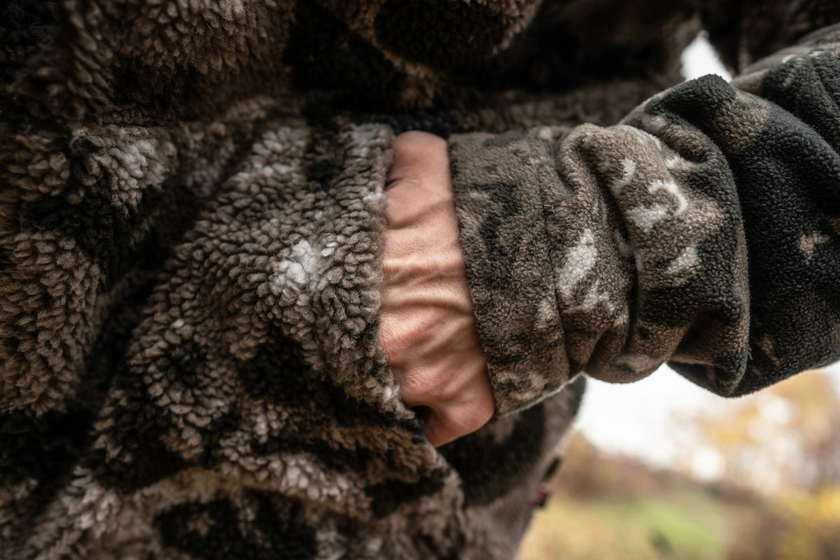 A person in a camouflage jacket holds the back of it, showcasing Pnuma Outdoors winter wear for hunters.