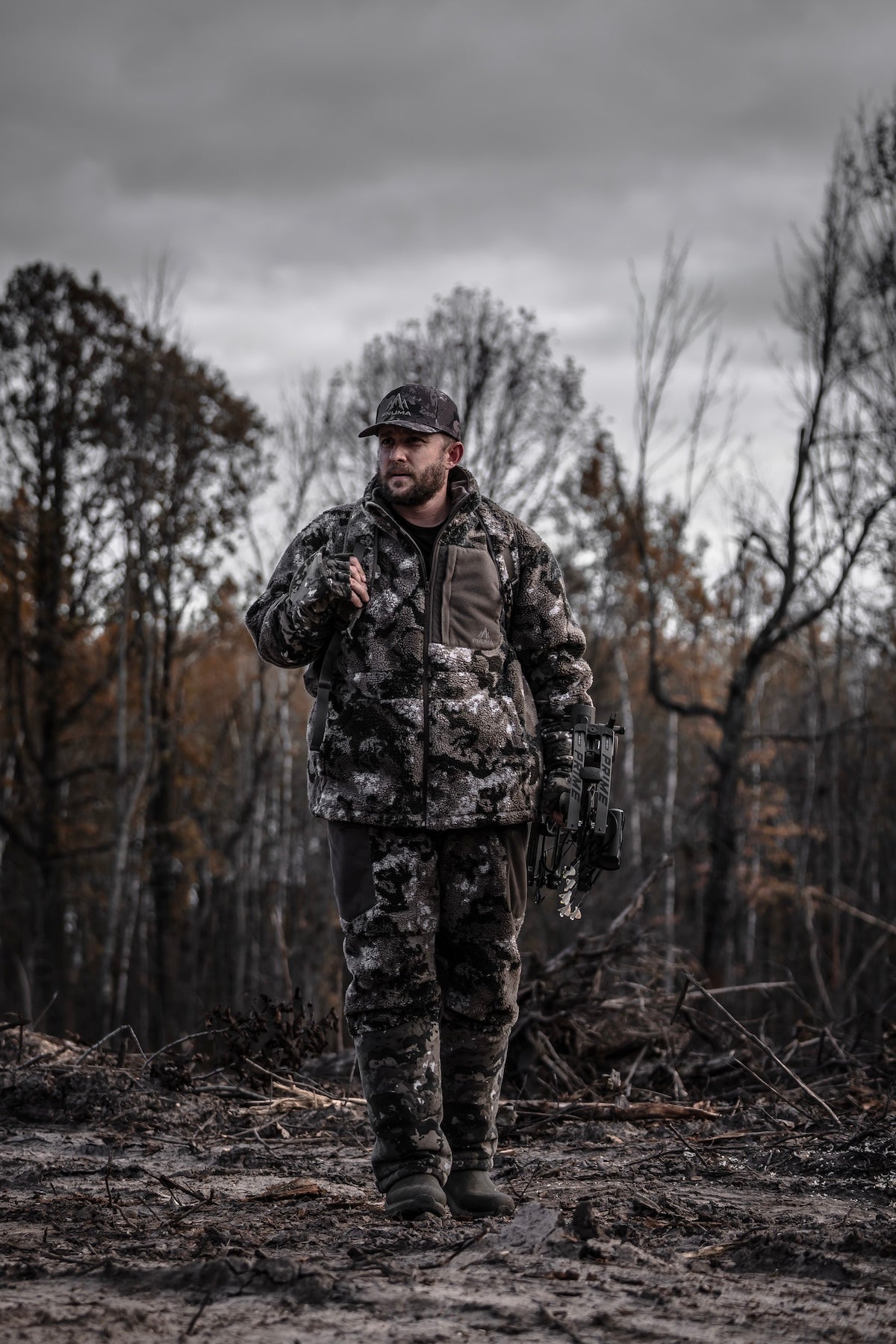 A man dressed in camouflage attire stands in a wooded area, surrounded by tall trees and foliage.