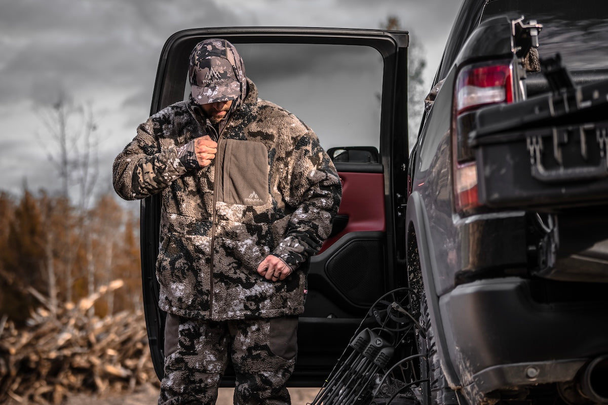A man in camouflage winter clothing is getting into a truck, representing Pnuma Outdoors' winter wear for outdoor adventures.