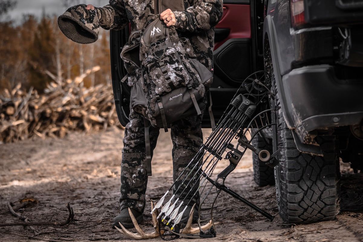 A man in camouflage winter gear stands beside his truck, showcasing Pnuma Outdoors winter wear for outdoor activities.