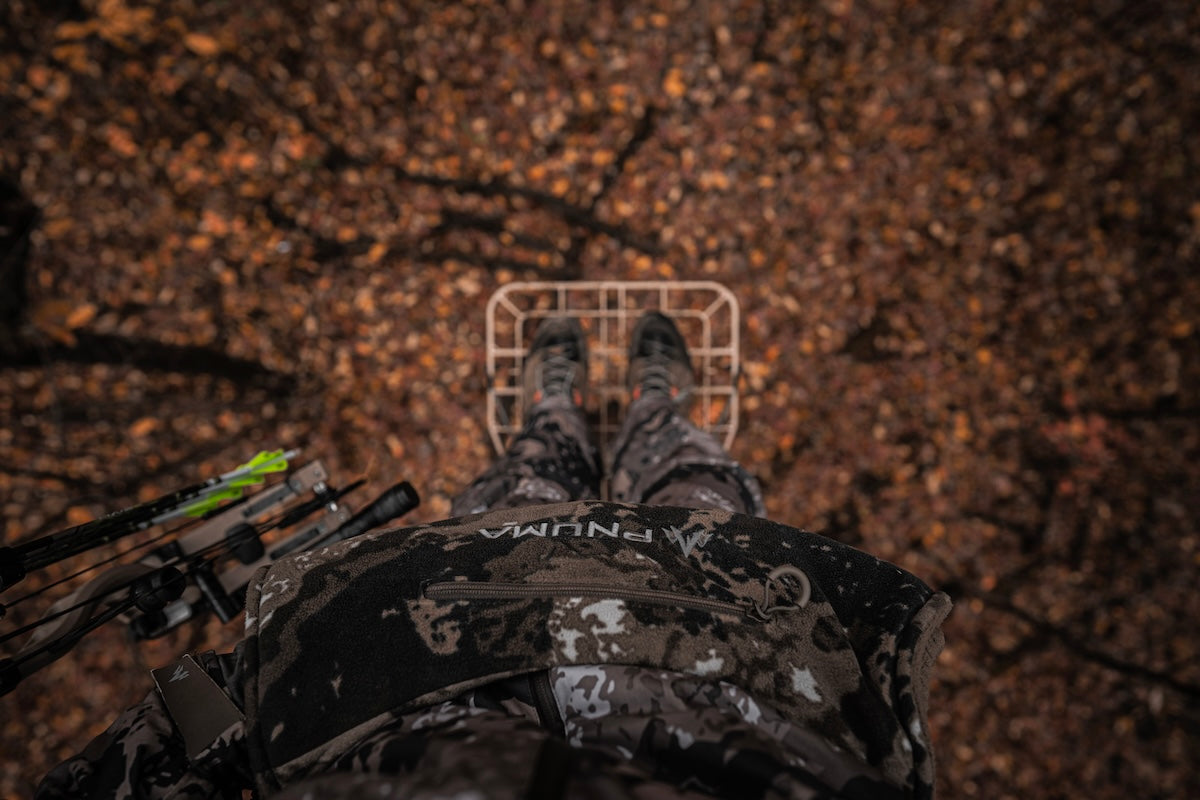 A person stands on a tree stump, holding a bow and arrow, showcasing Pnuma outdoor winter wear.