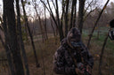 Hunter in camouflage standing in a wooded area at dusk, preparing gear while partially hidden among trees