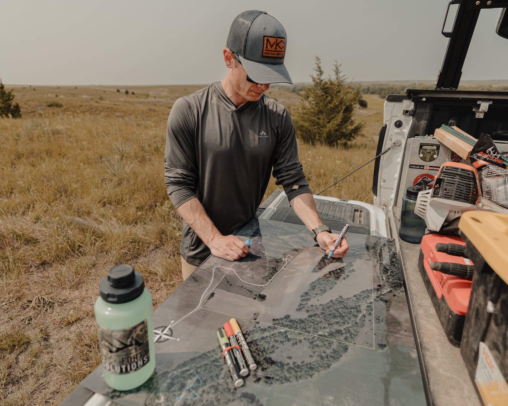 Man planning a route using a map spread on a truck bed, with gear and markers in an open grassland area.