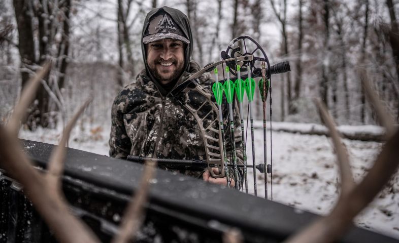 Smiling hunter in camouflage holding a compound bow, standing near antlers in a snowy forest.