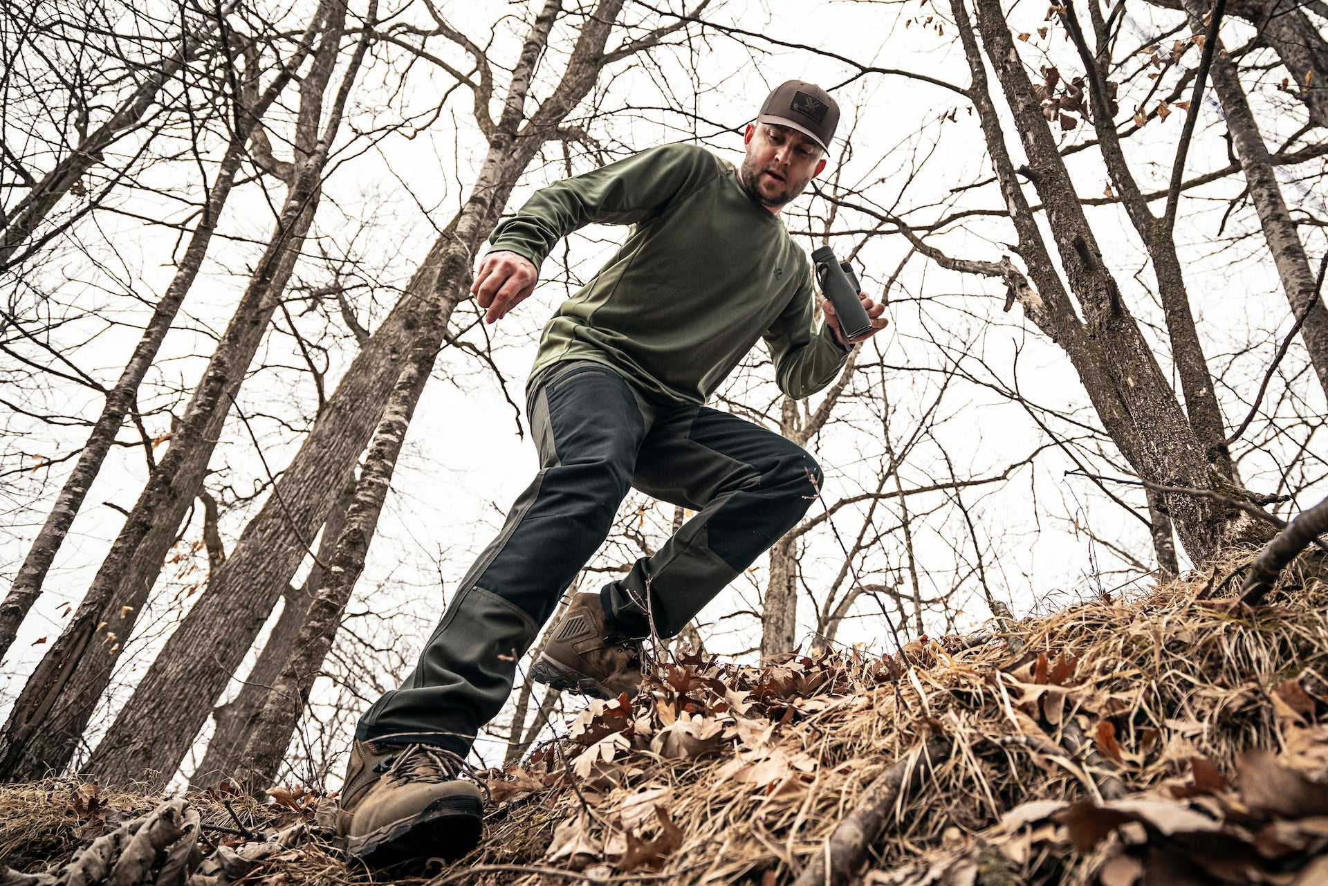 A man in a green shirt runs through a dense forest, surrounded by trees and foliage.