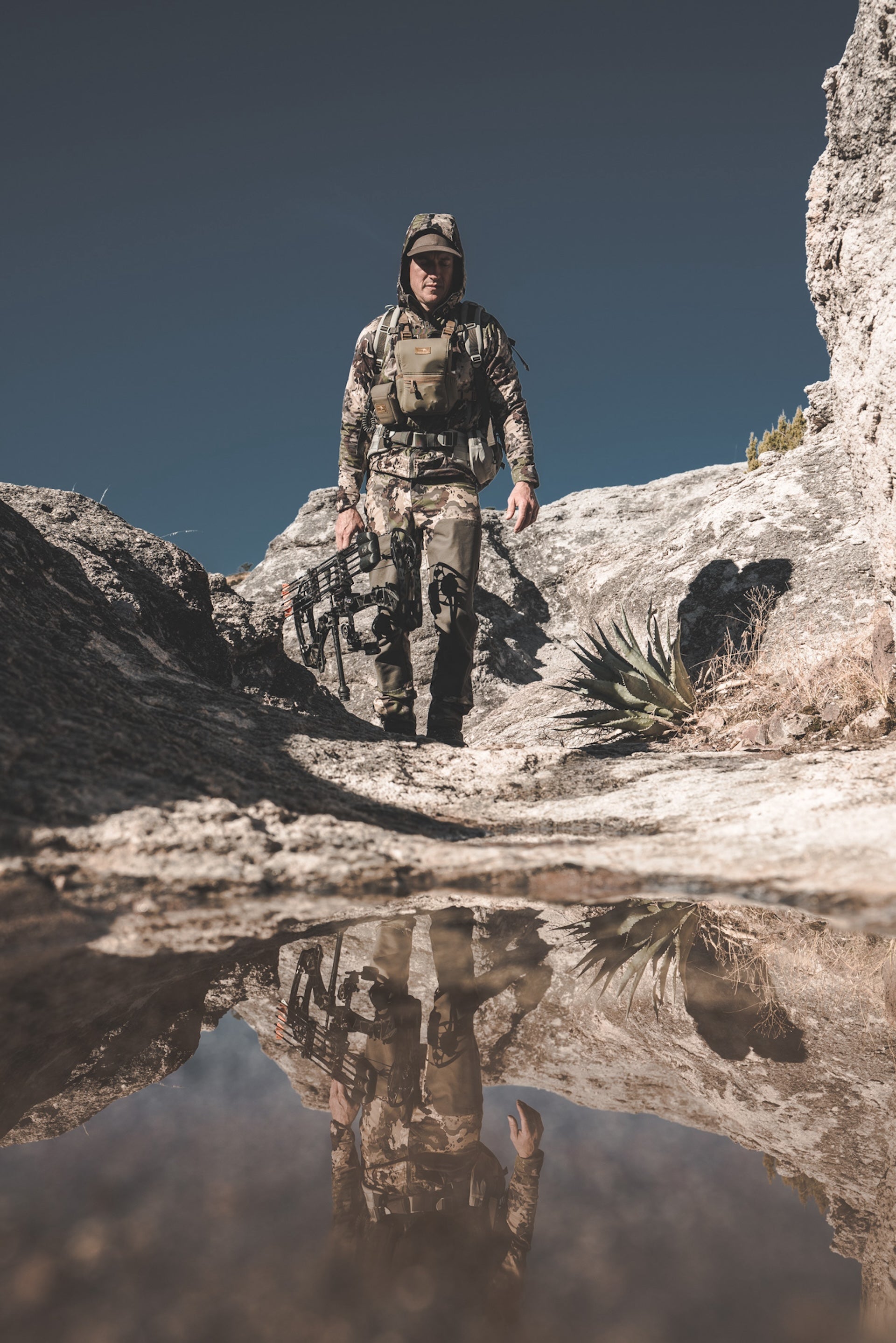 A soldier in camouflage stands on a rocky hill, surveying the landscape around him.