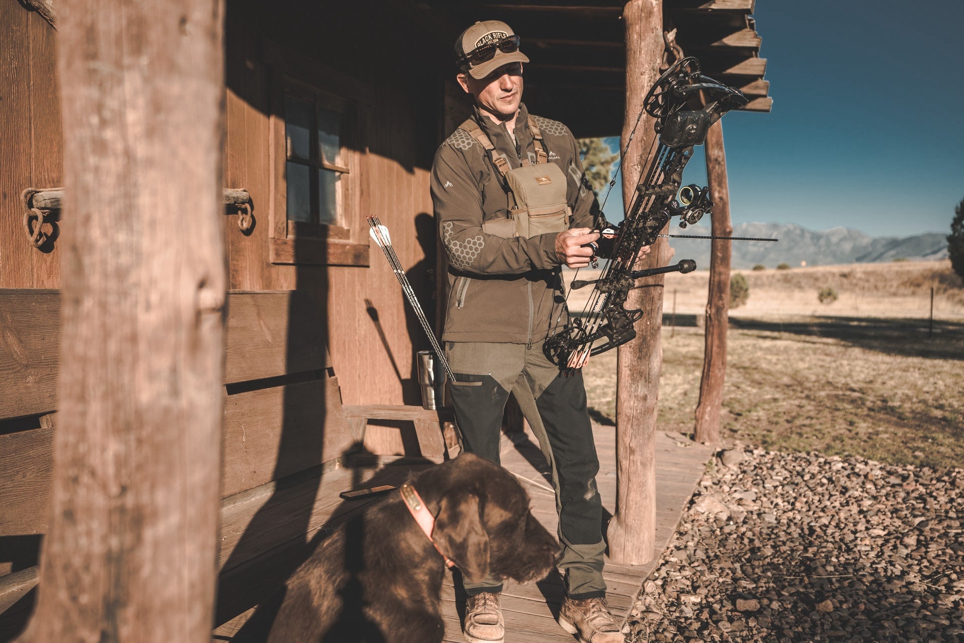 A man with a bow stands beside a dog outside a rustic cabin in a forested area.