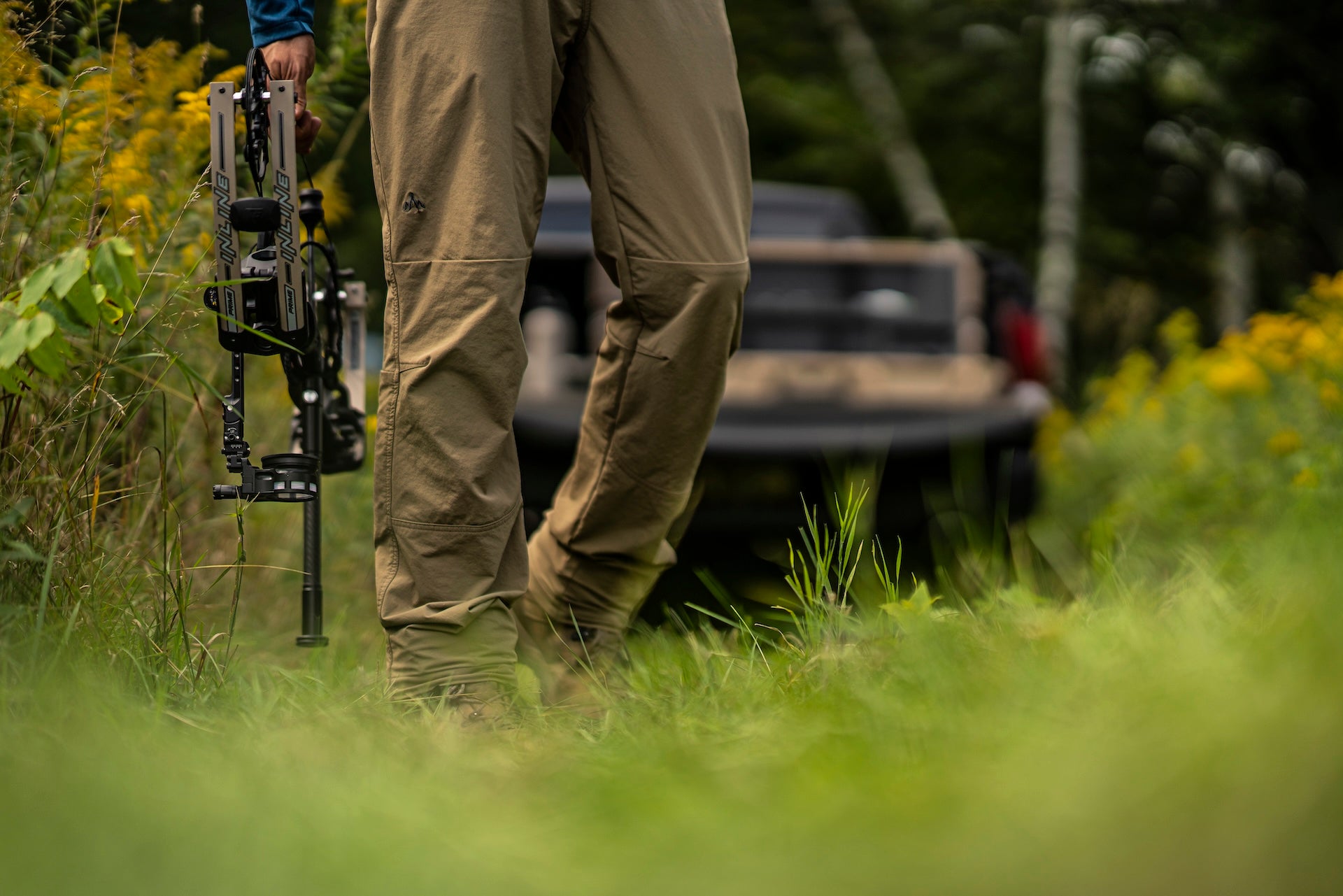 A man with a rifle stands in a grassy field, wearing camouflage clothing and looking intently into the distance.