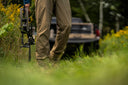 A man with a rifle stands in a grassy field, wearing camouflage clothing and looking intently into the distance.