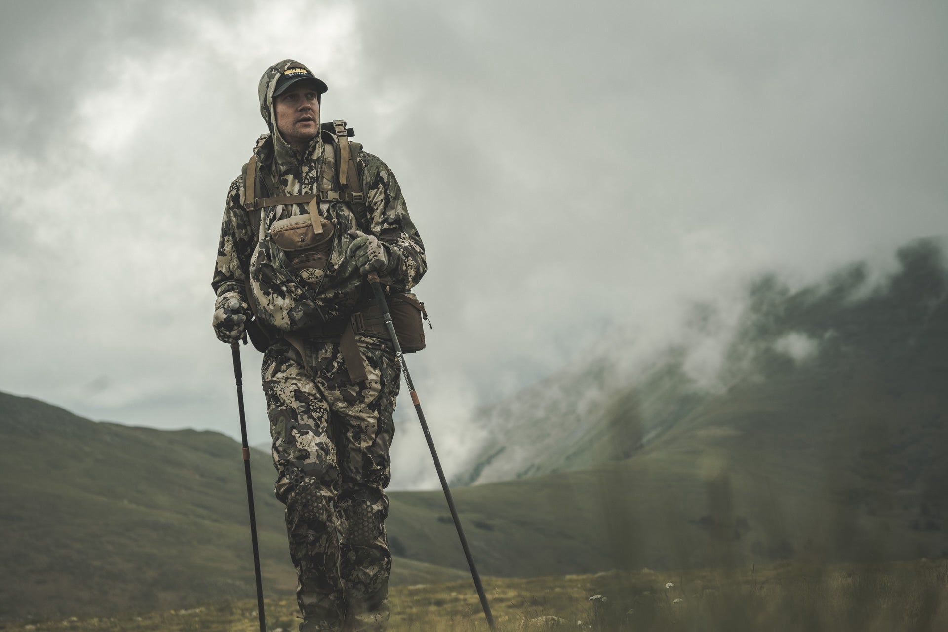Hunter in full camouflage gear hiking with trekking poles through a foggy mountain landscape